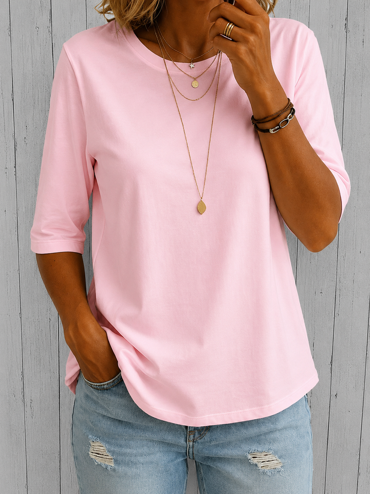 T-shirt rose pâle à demi-manches porté avec un jean clair et des colliers dorés, devant un fond de lattes grises. 5