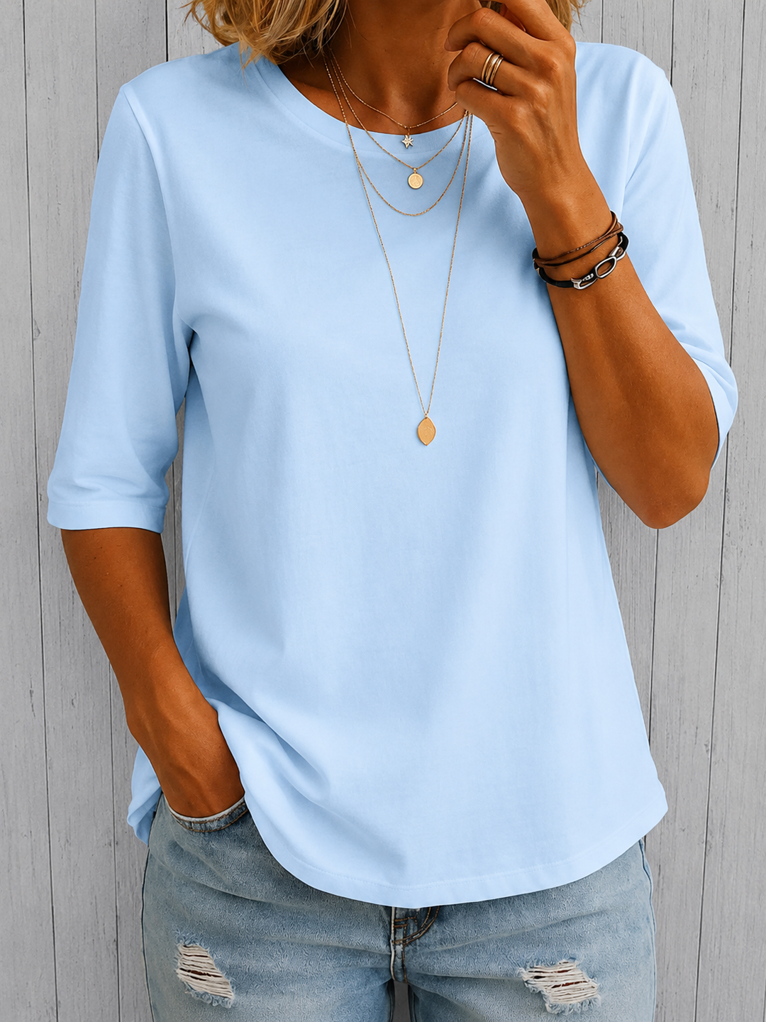 T-shirt bleu ciel à demi-manches porté avec un jean délavé et trois colliers dorés, devant un mur en bois gris. 3