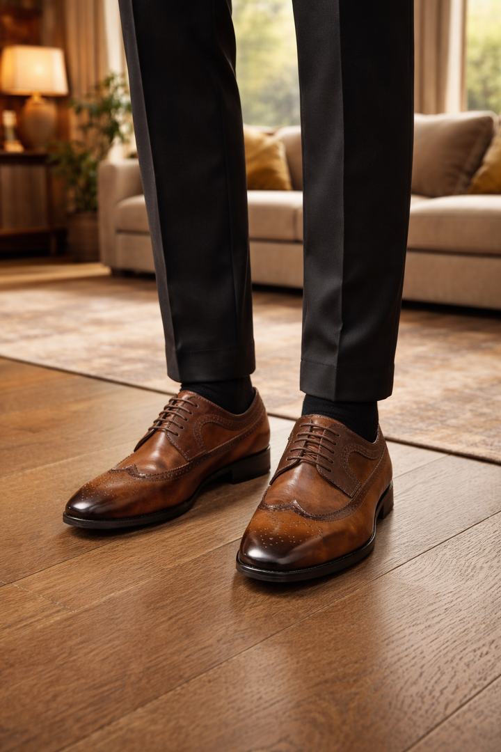 Homme debout en pantalon noir portant les chaussures en cuir brun à lacets et perforations, sur un parquet de salon. 1