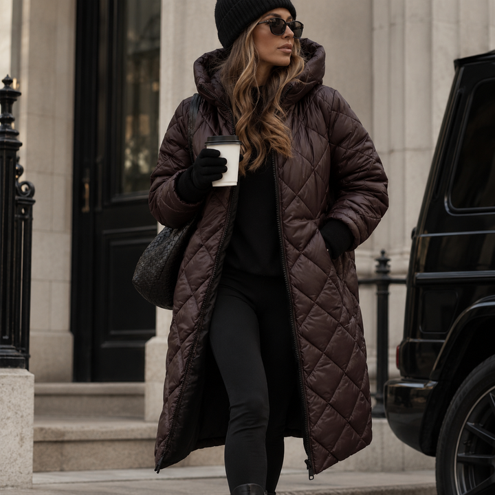 Femme portant le manteau long matelassé à capuche brun foncé, zip ouvert, avec bonnet noir, lunettes de soleil et sac tressé, devant un porche. 2