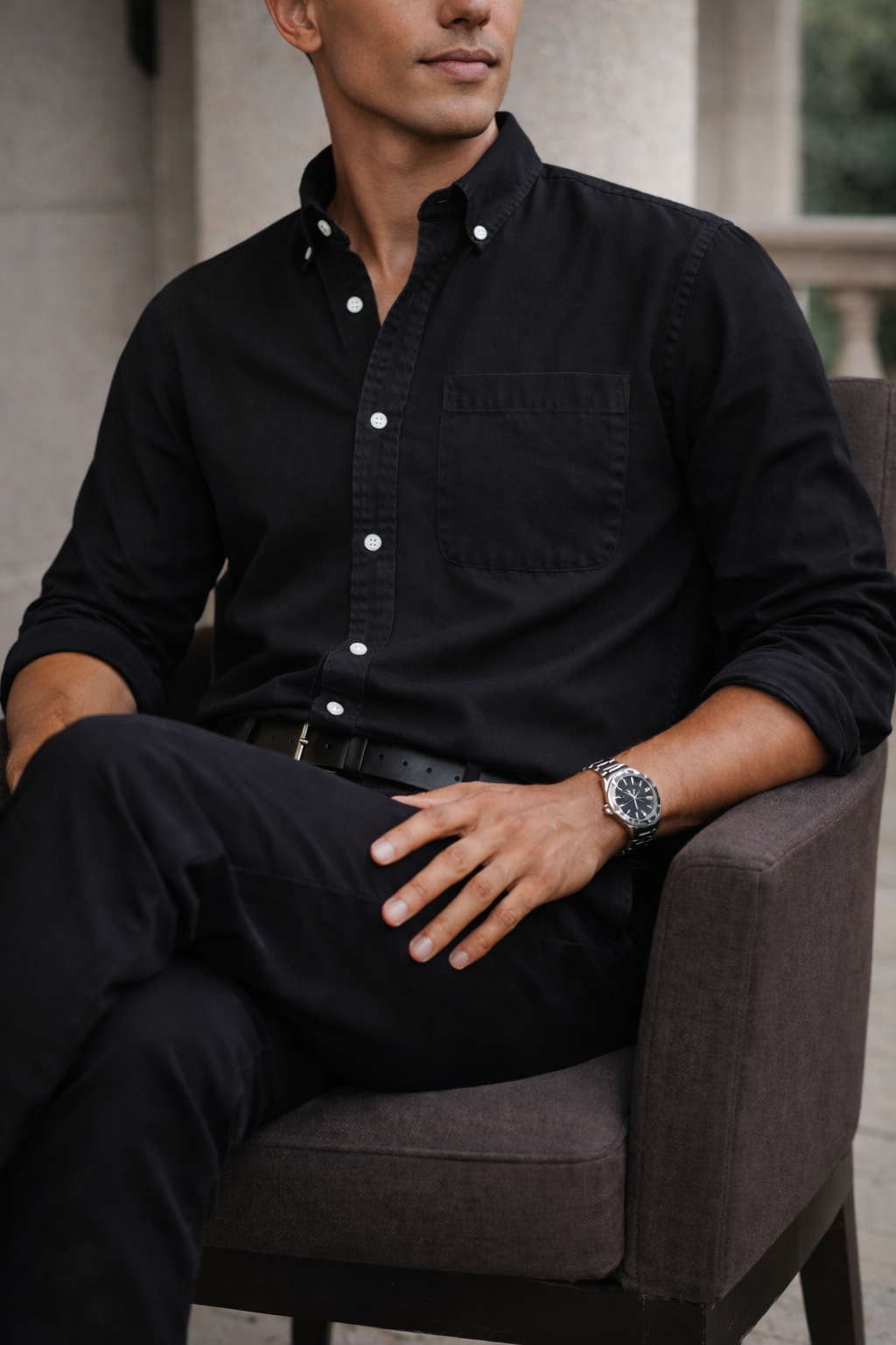 Chemise noire à col boutonné portée avec un pantalon noir, le modèle assis dans un fauteuil gris foncé. 7