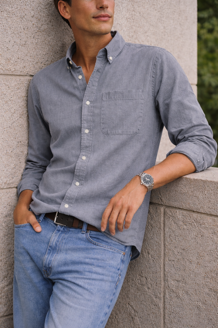 Chemise grise à col boutonné portée avec un jean bleu clair, le modèle adossé à un mur en pierre. 5