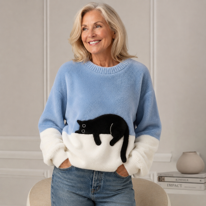 Femme blonde souriante portant le pull bleu clair et blanc en maille duveteuse avec un chat noir allongé, mains dans les poches. 1
