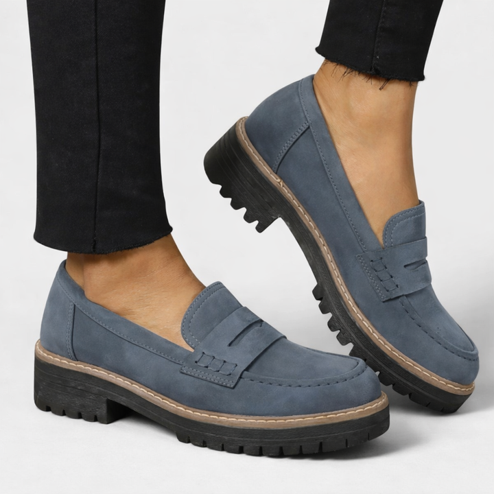 Mocassins bleu gris en aspect daim portés avec un pantalon noir, l'un posé et l'autre levé montrant la semelle crantée. 3