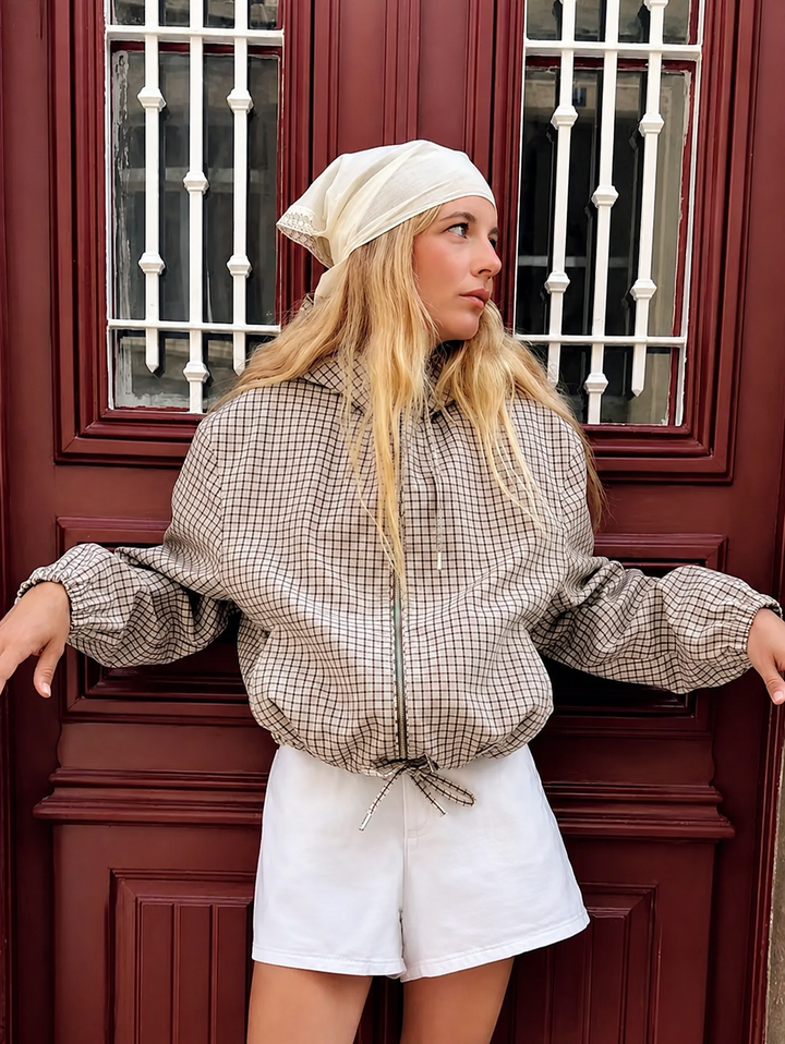 Mannequin en plan rapproché devant une porte rouge, veste à petits carreaux portée avec un foulard crème et un short blanc, bras écartés. 1