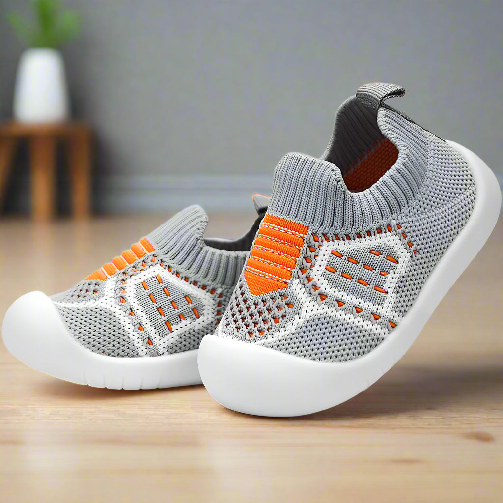 Chaussures en maille grise à rayures orange, motif blanc et semelle blanche souple, présentées en angle sur un parquet clair. 16