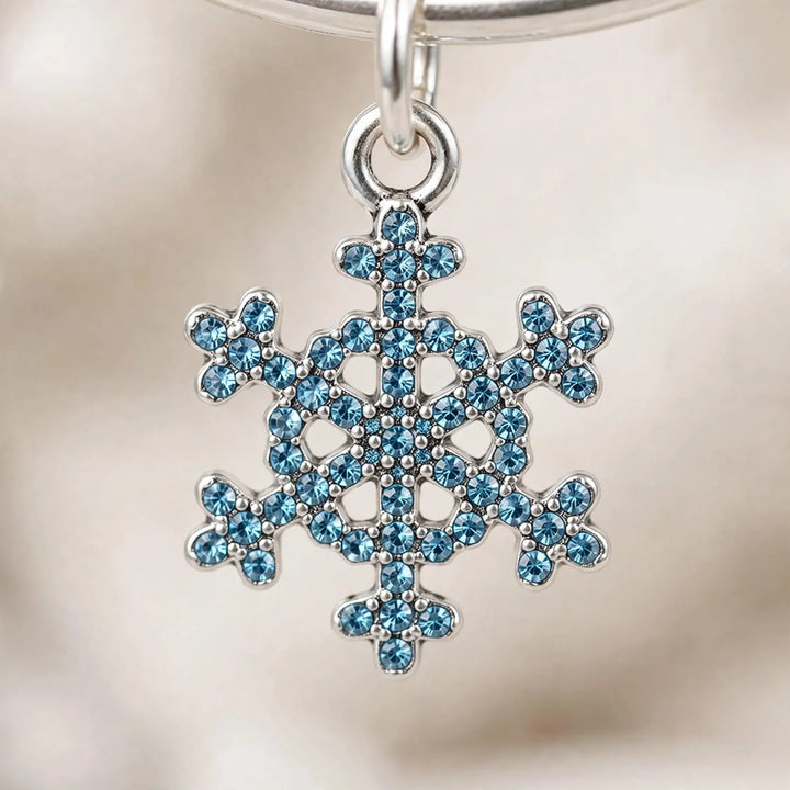 Gros plan sur un charm flocon de neige argenté serti de strass bleus, suspendu à un anneau 3