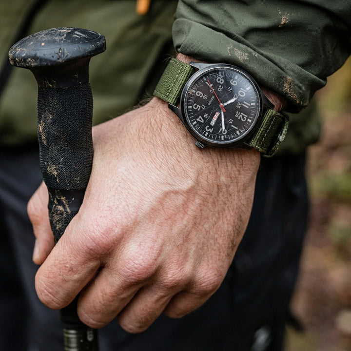 Montre portée au poignet d'un randonneur tenant un bâton de marche, manche de veste verte tachée de boue. 2