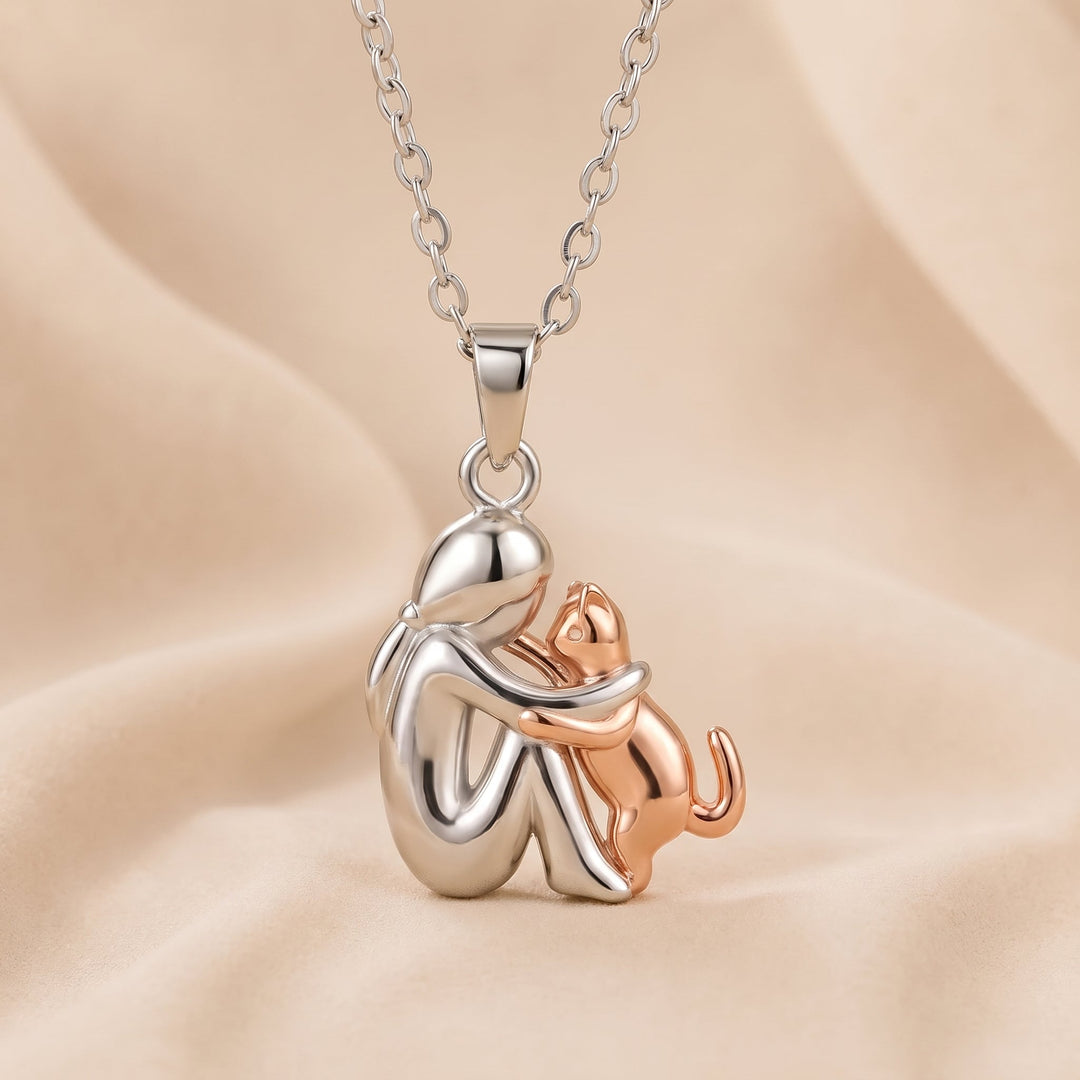 Pendentif d'une personne argentée brillante enlacée par un chat couleur cuivre rosé, posé sur un drap beige ondulé. 4