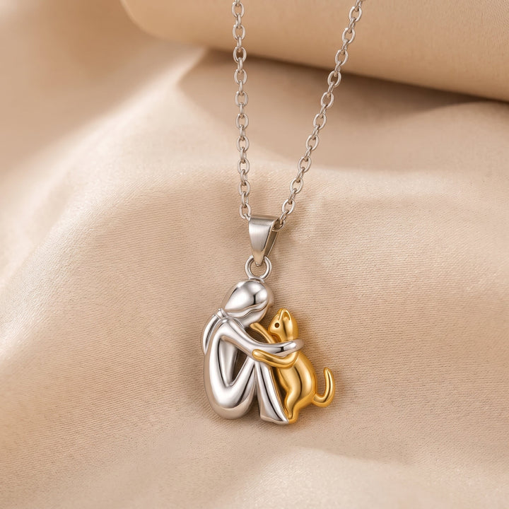 Pendentif brillant d'une personne argentée assise enlaçant un chat doré, présenté en légère plongée sur un tissu satiné beige. 2