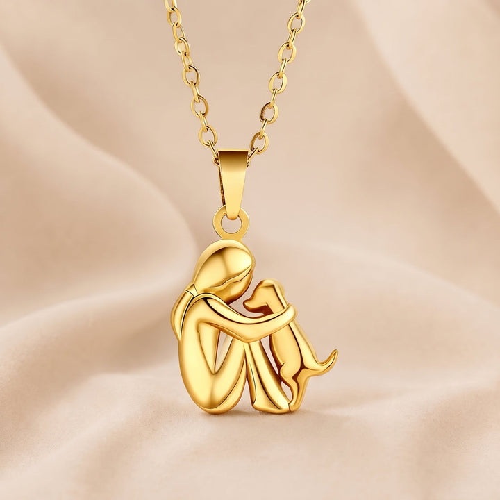 Pendentif entièrement doré et brillant, une personne assise entourant un chien de son bras, avec chaîne dorée sur tissu beige. 5