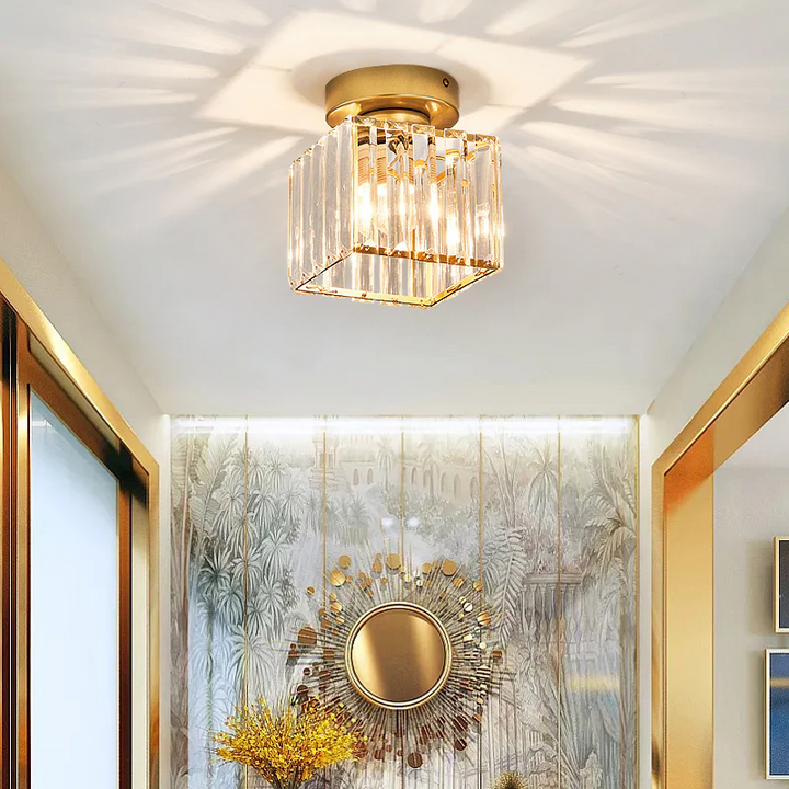 Plafonnier carré en cristal à finition dorée allumé dans un couloir, face à un miroir soleil doré et un mur à motif. 9