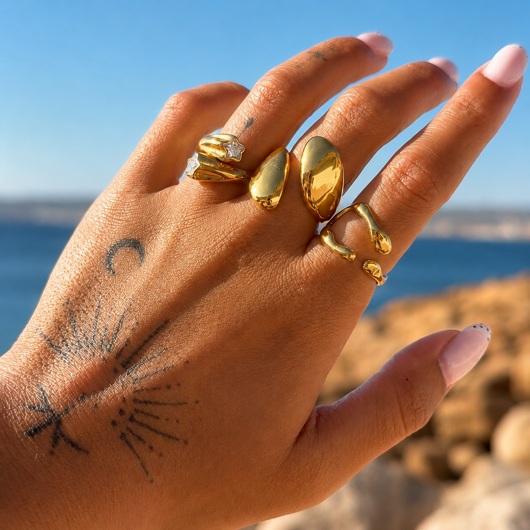 Main tatouée portée devant la mer, avec la bague dorée ouverte à l'index parmi plusieurs autres bagues dorées. 3