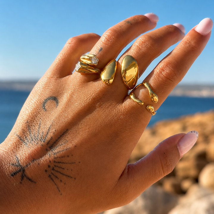 Main tatouée portée devant la mer, avec la bague dorée ouverte à l'index parmi plusieurs autres bagues dorées. 3