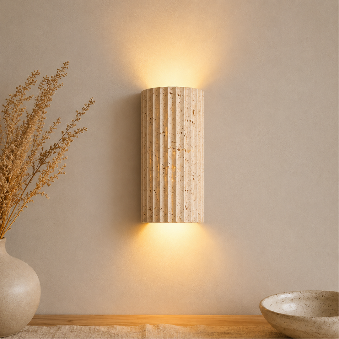 Applique cannelée en pierre beige allumée sur un mur clair, diffusant une lumière chaude vers le haut et le bas, près d'un vase. 1