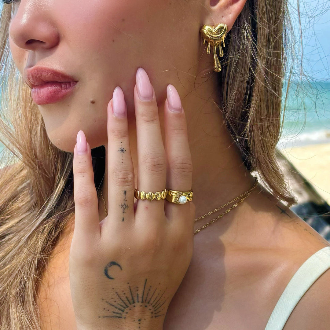 Gros plan sur une femme portant la boucle d'oreille cœur fondant dorée, avec des bagues dorées, sur fond de plage. 4