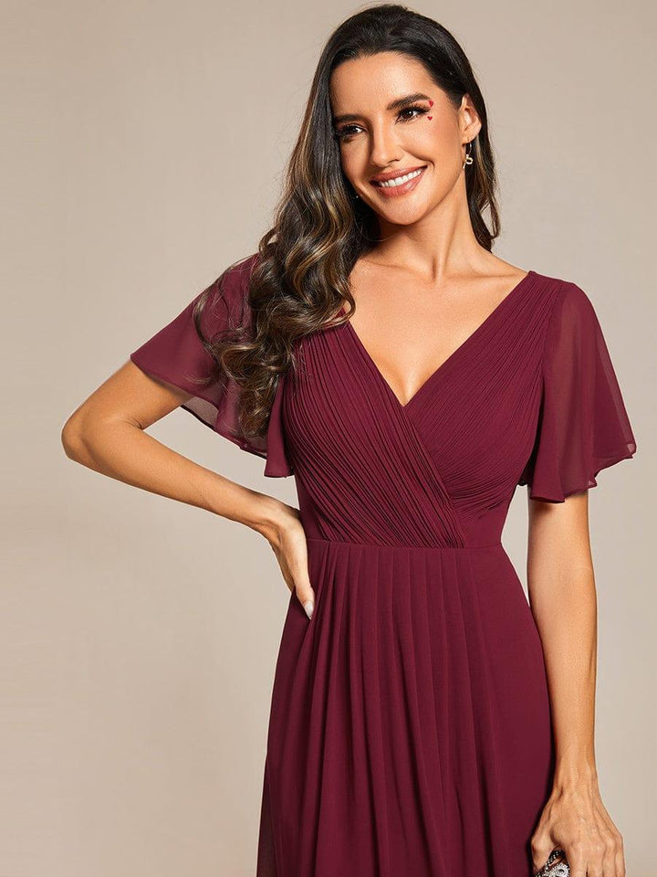 Gros plan sur le décolleté en V plissé croisé et les manches volantées de la robe bordeaux, mannequin souriante main sur la hanche. 46