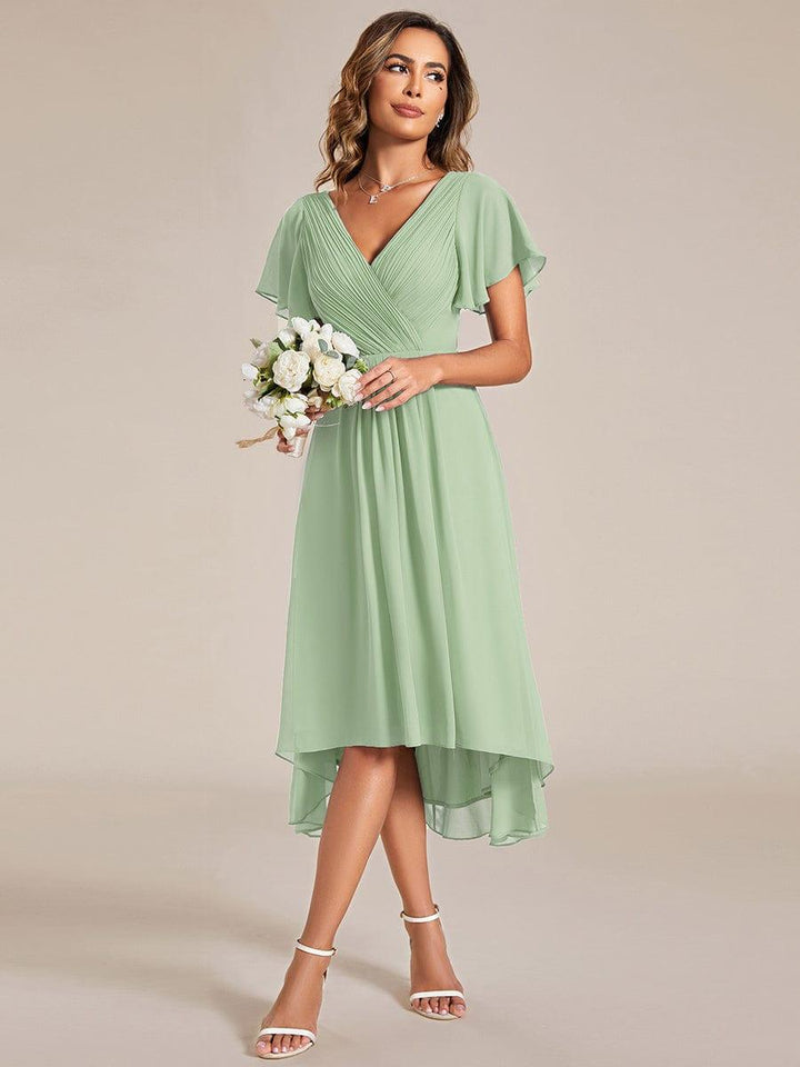 Robe vert sauge portée par une mannequin tenant un petit bouquet de fleurs blanches, col en V plissé et jupe asymétrique. 79