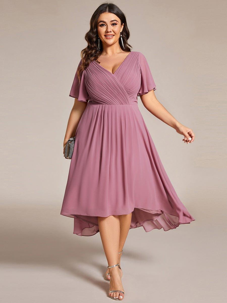Robe mauve rosé portée de face par une mannequin en grande taille, tenant une pochette argentée, jupe fluide à ourlet asymétrique. 63