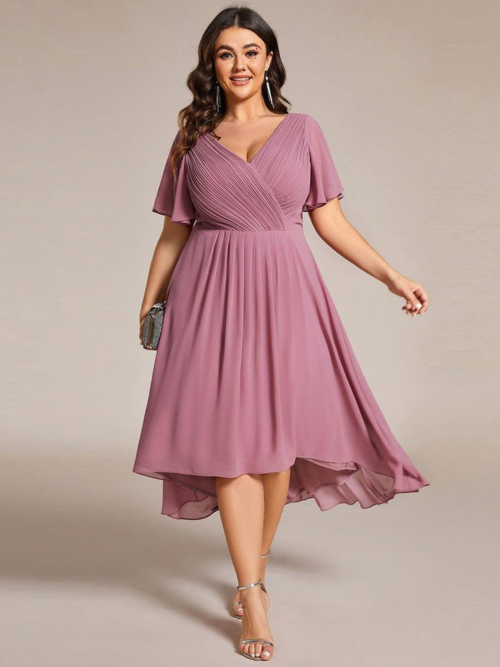Robe mauve rosé portée de face par une mannequin en grande taille, tenant une pochette argentée, jupe fluide à ourlet asymétrique. 63