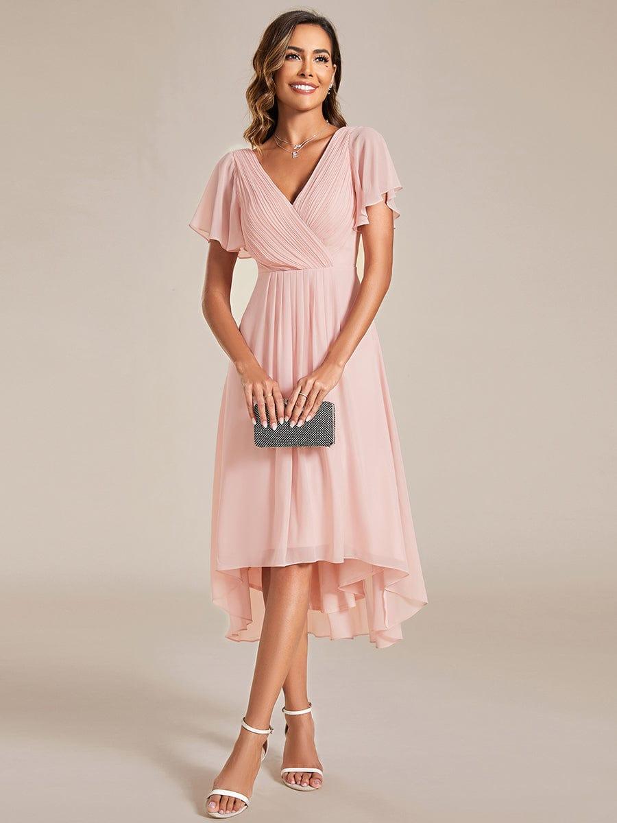 Robe rose pâle portée de face, pochette noire scintillante tenue à deux mains, buste plissé et ourlet asymétrique. 15