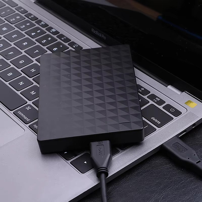 Disque dur noir posé sur le clavier d'un ordinateur portable, relié par un câble USB noir à un port de la machine. 3