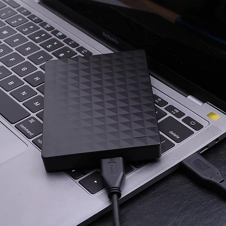 Disque dur noir posé sur le clavier d'un ordinateur portable, relié par un câble USB noir à un port de la machine. 3