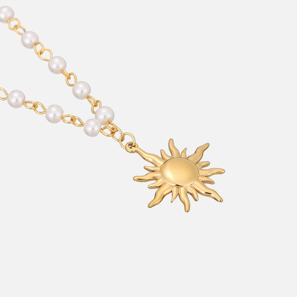 Gros plan en vue inclinée sur le pendentif soleil doré et les perles blanches de la chaîne, posés sur fond gris clair. 2