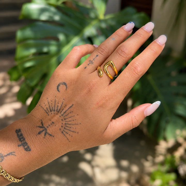 Anneau doré porté à l'index d'une main tatouée, devant un feuillage vert en plein soleil 3