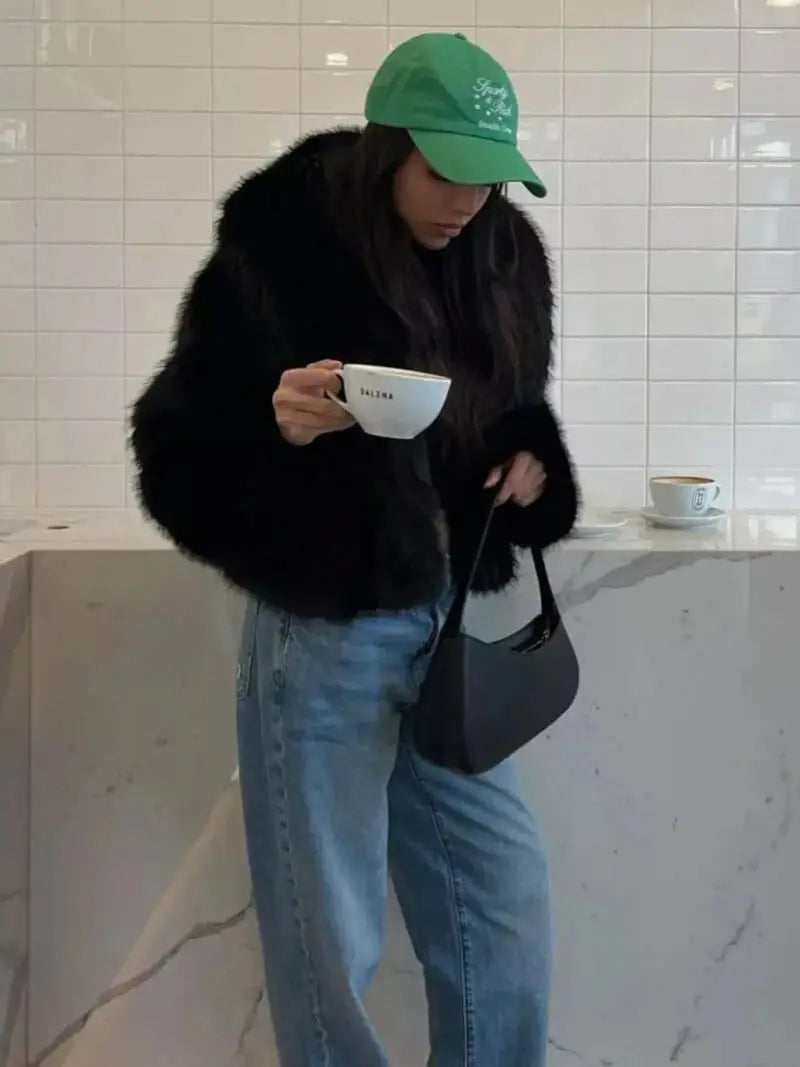 Femme en casquette verte tenant une tasse de café, vêtue du manteau noir en fausse fourrure et d'un jean 10