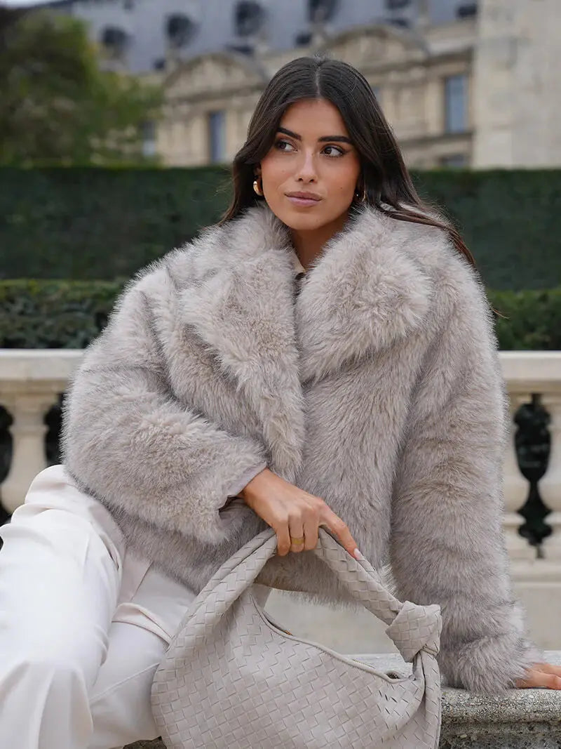 Femme assise en extérieur portant le manteau en fausse fourrure gris clair, un sac tressé beige à la main 6