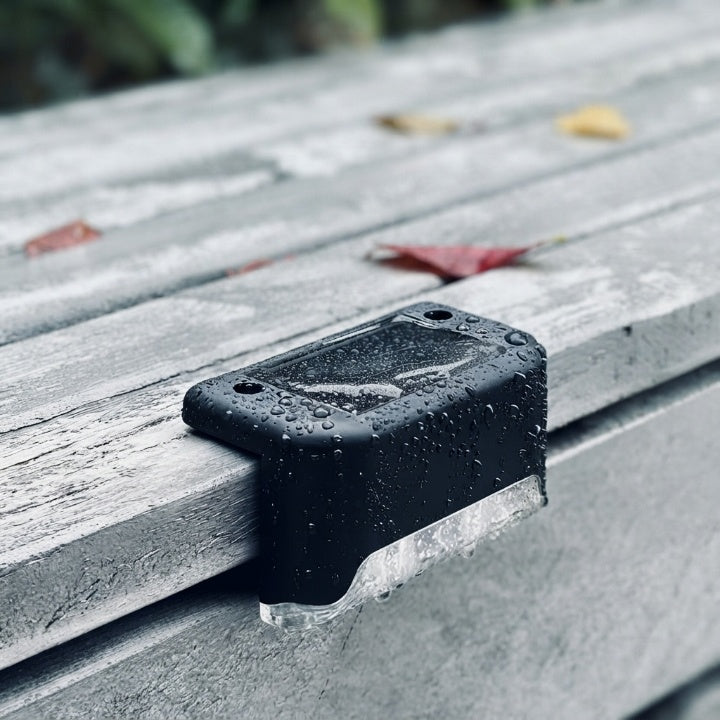 Gros plan d'une lampe solaire noire couverte de gouttes de pluie sur une planche en bois grisé, avec feuilles d'automne autour. 7