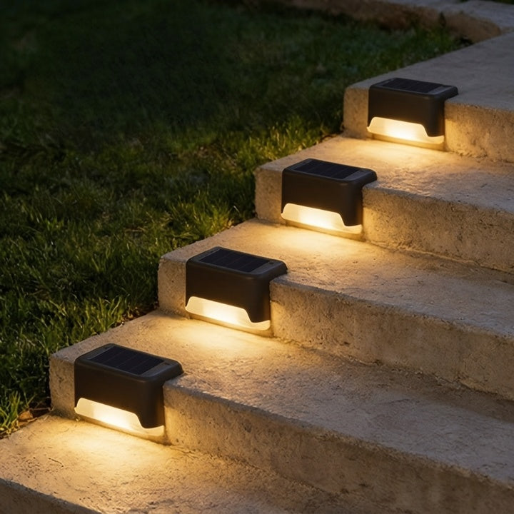 Quatre lampes solaires noires alignées sur un escalier en pierre au bord d'une pelouse, avec panneau solaire visible et lueur chaude. 3