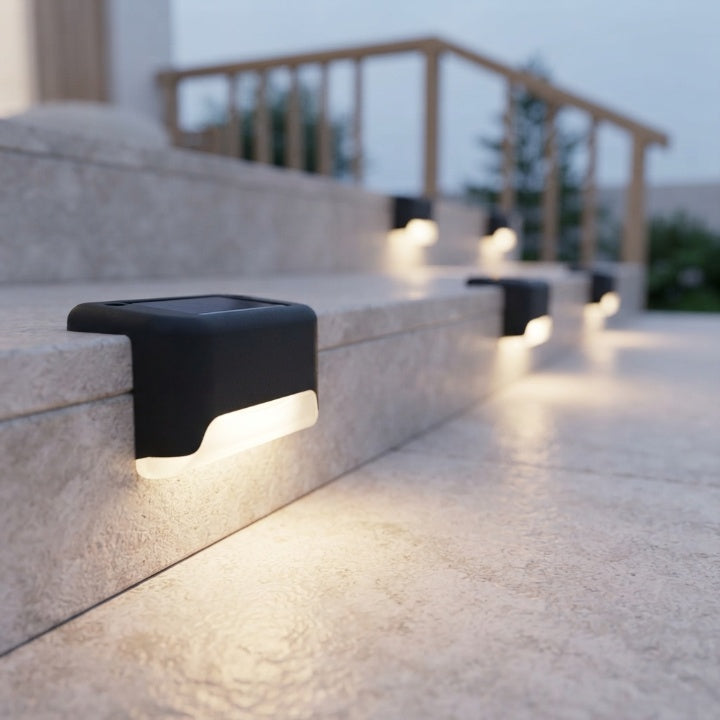 Lampes solaires noires fixées au bord de marches en pierre claire, diffusant une lumière chaude au crépuscule devant une rambarde en bois. 1