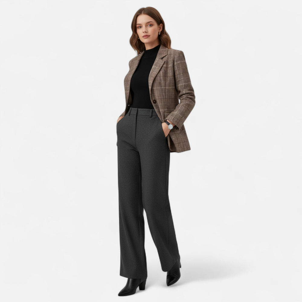 Mannequin en pied portant le blazer à carreaux avec un haut noir, un pantalon gris foncé et des bottines noires, mains dans les poches. 2