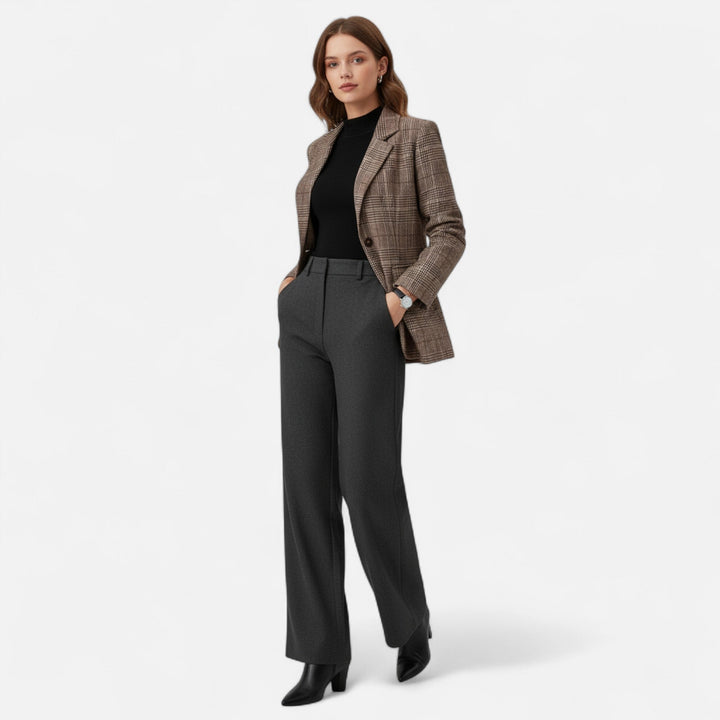 Mannequin en pied portant le blazer à carreaux avec un haut noir, un pantalon gris foncé et des bottines noires, mains dans les poches. 2