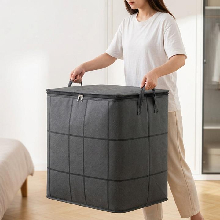Femme en t-shirt blanc portant à deux mains le grand sac gris fermé par ses anses, dans une chambre. 2