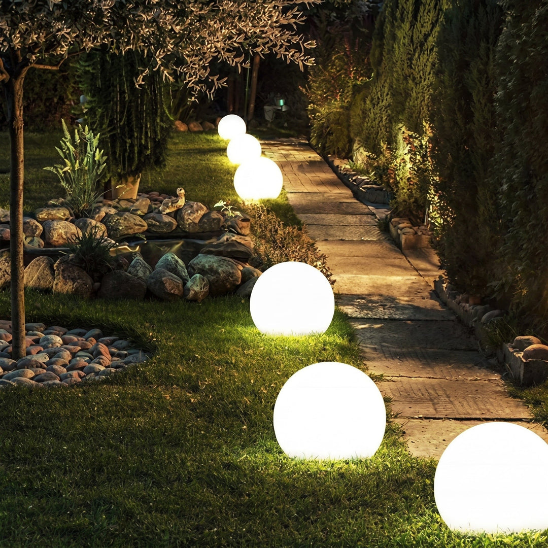Sphères lumineuses de tailles décroissantes alignées sur la pelouse le long d'une allée dallée, de nuit, près d'un bassin de pierres. 6