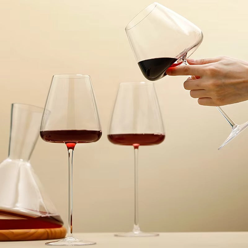Une main incline un verre de vin rouge au-dessus de deux verres posés, près d'une carafe 9