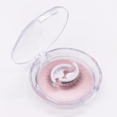 Boîtier rond transparent ouvert contenant une paire de faux cils sur un support blanc, posé sur un fond rose nacré. 1