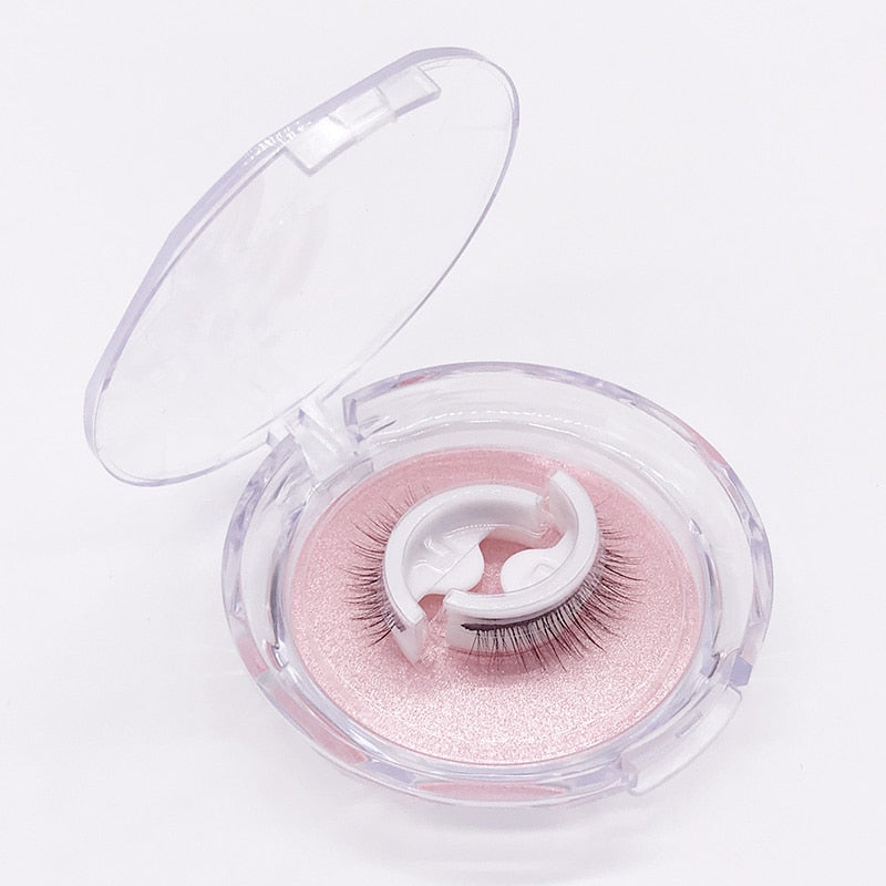 Boîtier rond transparent ouvert, fond blanc, laissant voir une paire de faux cils sur un support blanc posé sur un coussin rose nacré. 6