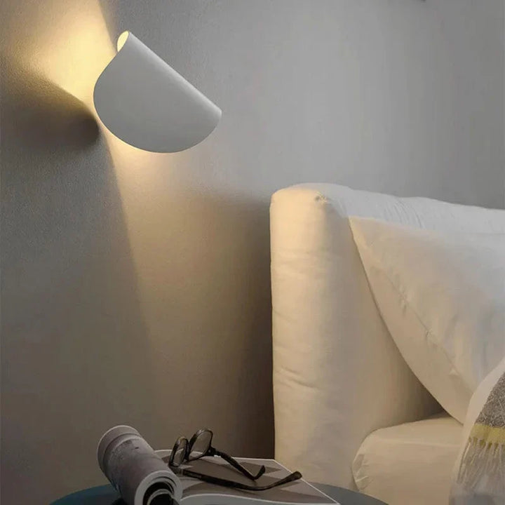 Applique blanche inclinée au-dessus d'un chevet, lumière chaude dirigée vers le haut, avec lunettes et magazine posés sur une table. 7
