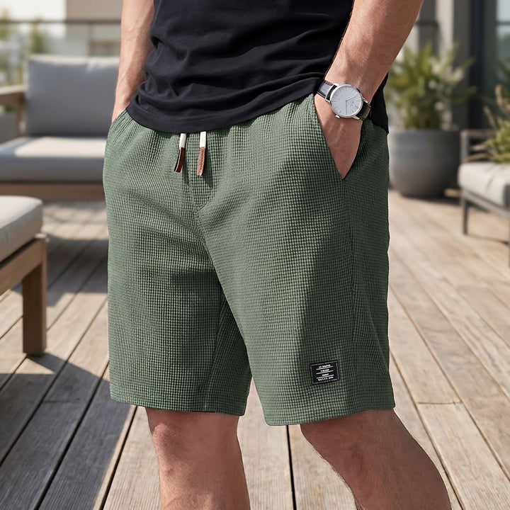 Short vert kaki en tissu gaufré à cordon de serrage, porté avec un tee-shirt noir et une montre sur une terrasse en bois. 5