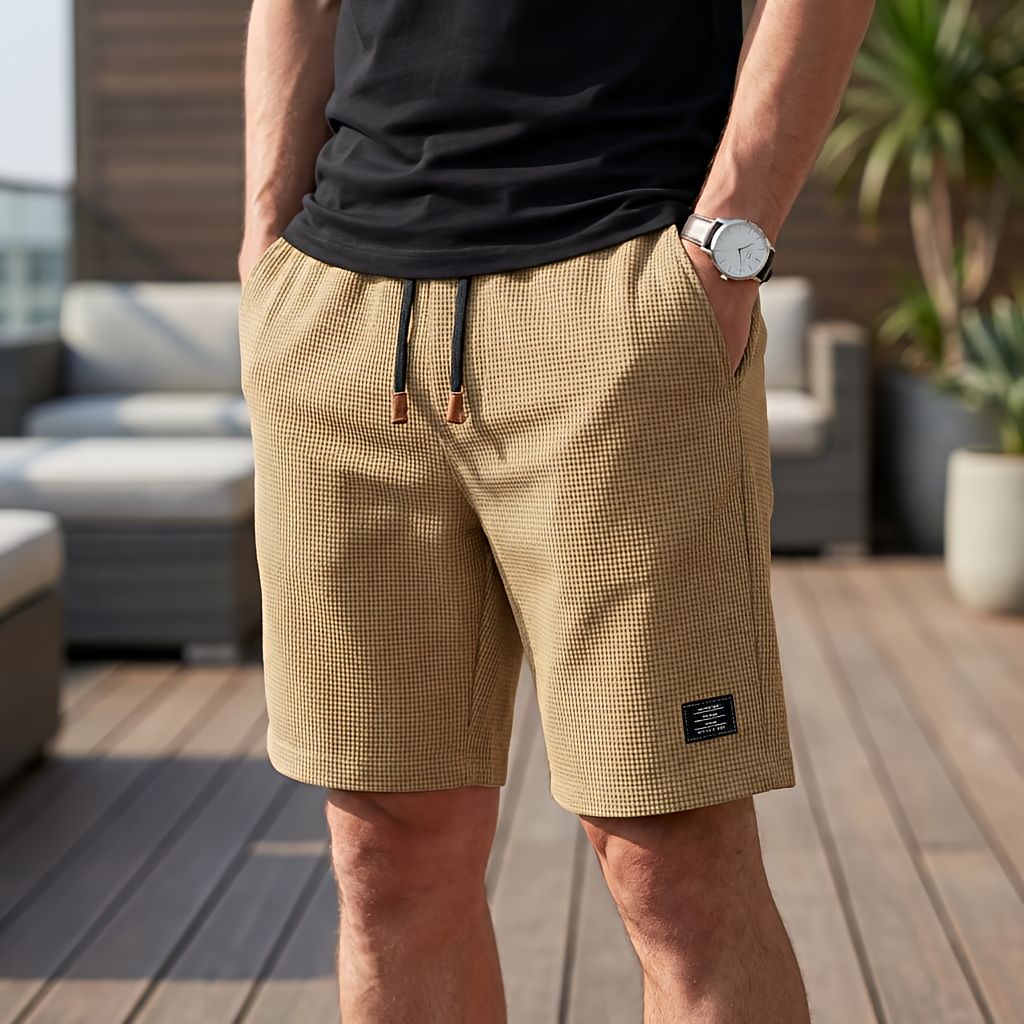Short beige foncé en tissu gaufré à cordon noir, porté avec un tee-shirt noir, une main dans la poche, sur une terrasse. 4