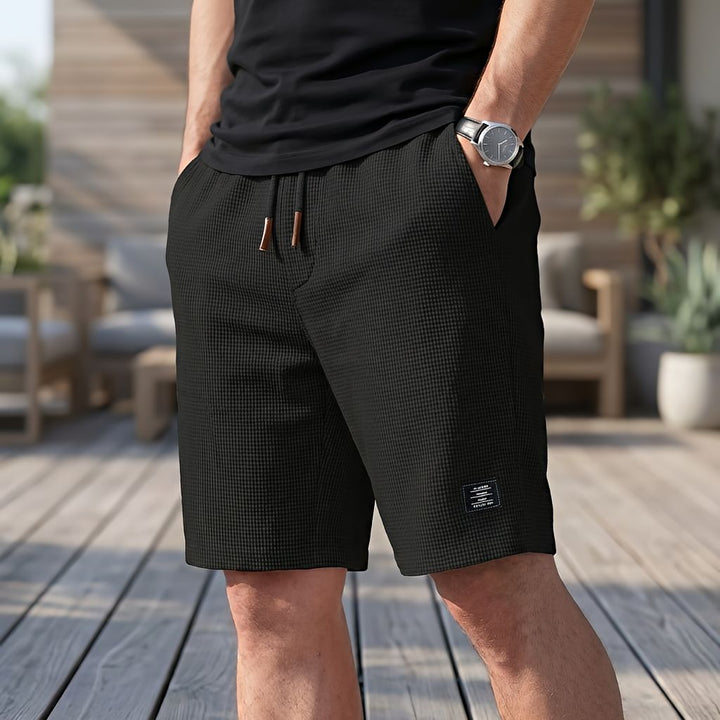 Short noir en tissu gaufré avec cordon de serrage, porté avec un tee-shirt noir et une montre, une main dans la poche. 3