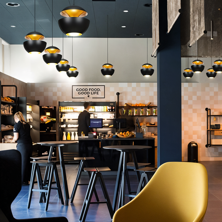 Nombreuses suspensions noires et dorГ©es de tailles variГ©es dans un cafГ©-boutique moderne avec tables hautes et fauteuil jaune. 4