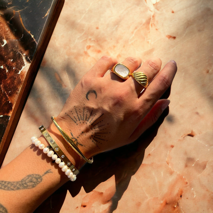 Main et poignet tatoués posés sur un marbre rosé, portant le jonc doré avec un bracelet de perles et deux bagues dorées. 4