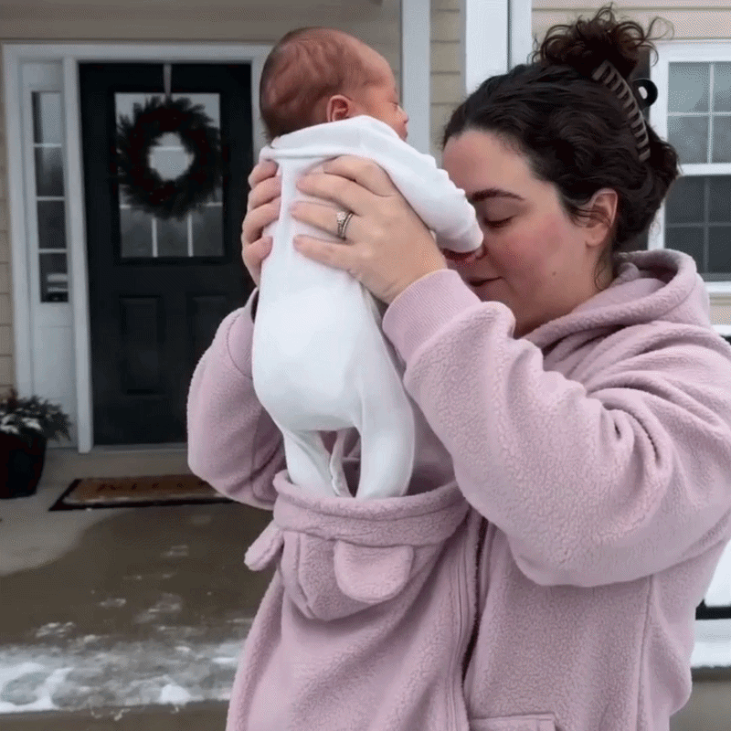 Femme en sweat rose soulevant un nouveau-né vêtu de blanc hors de l'empiècement porte-bébé, devant une porte d'entrée. 11