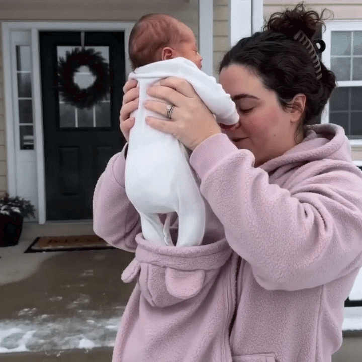 Femme en sweat rose soulevant un nouveau-né vêtu de blanc hors de l'empiècement porte-bébé, devant une porte d'entrée. 11