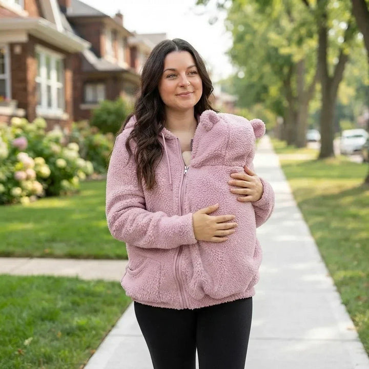 Femme marchant sur un trottoir en sweat rose zippé, la main sur l'empiècement porte-bébé à oreilles d'ourson. 15